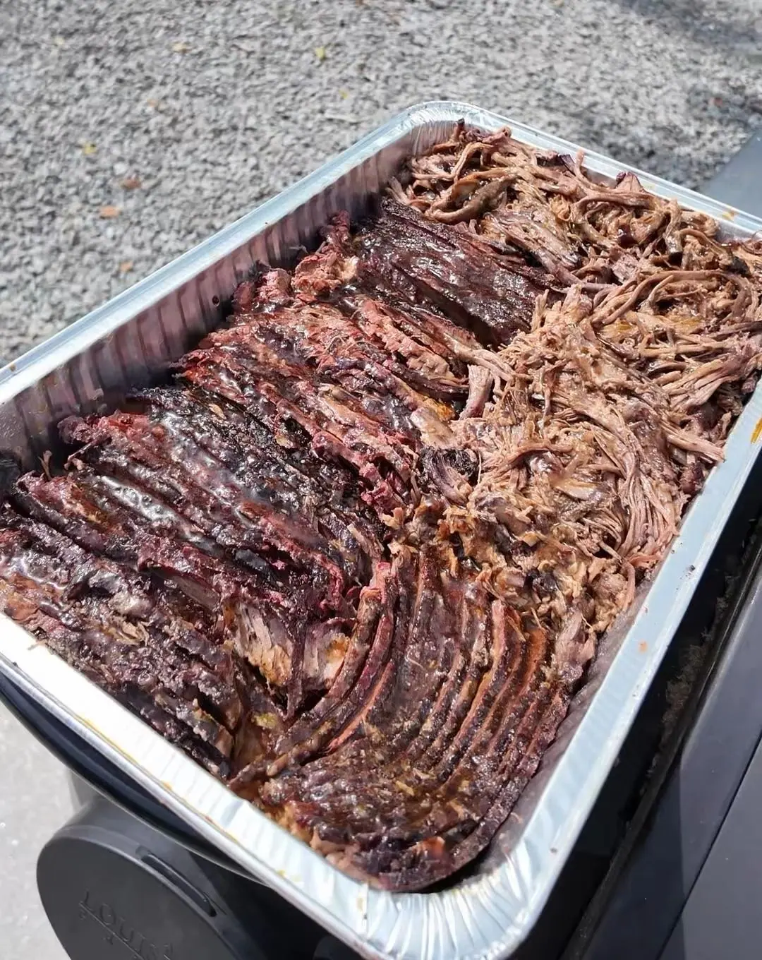 A full foil tray of sliced and pulled brisket set on the smoker outside, gravel and grass behind