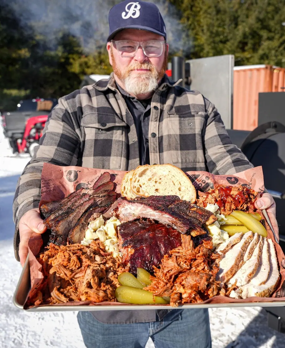 Aaron holding a loaded sheet tray of brisket, ribs, pulled pork, sliced chicken, pickles and bread beside the black smoker with its round red Barrie’s Best BBQ badge