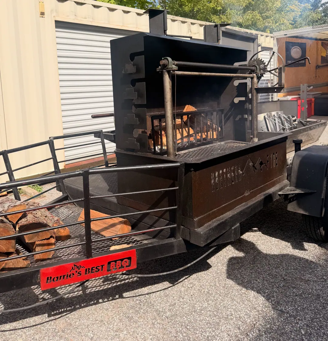 The open-fire grill trailer: logs burning in the fire basket, grates and crank above, firewood in the rack and the red Barrie’s Best BBQ plate