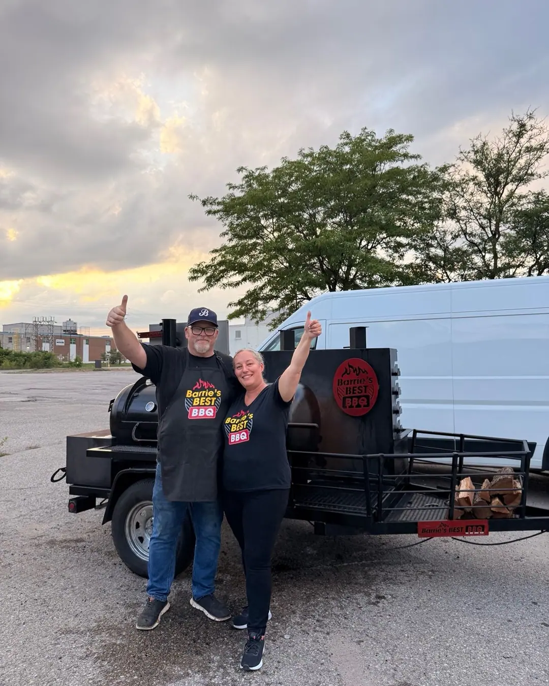 Jenn and Aaron in Barrie’s Best BBQ shirts giving a thumbs up beside their smoker trailer at sunset