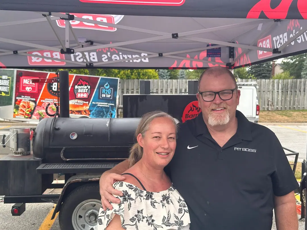 Jenn and Aaron smiling under the Barrie’s Best BBQ tent, the smoker trailer beside them