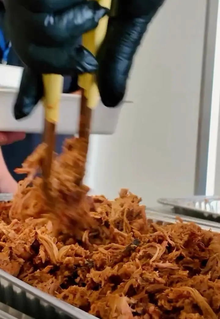 Gloved hands lifting a tangle of pulled pork with tongs from a full chafing pan