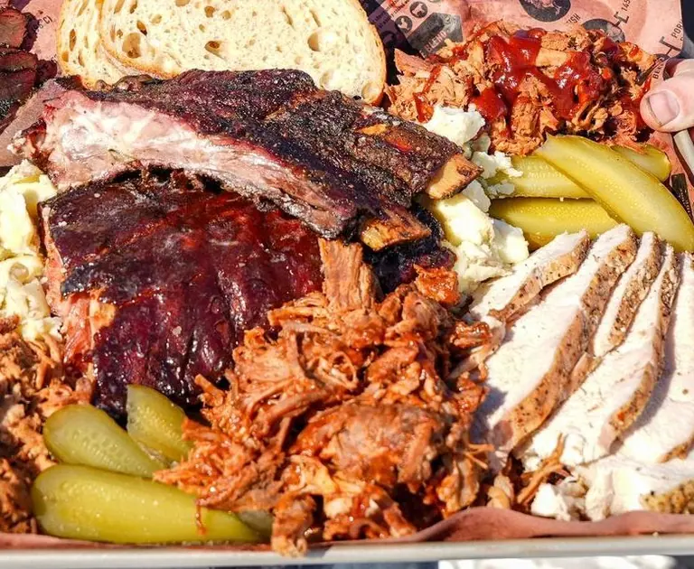Close up of sauced pork ribs with dark bark, pulled pork, sliced smoked chicken and pickles on butcher paper