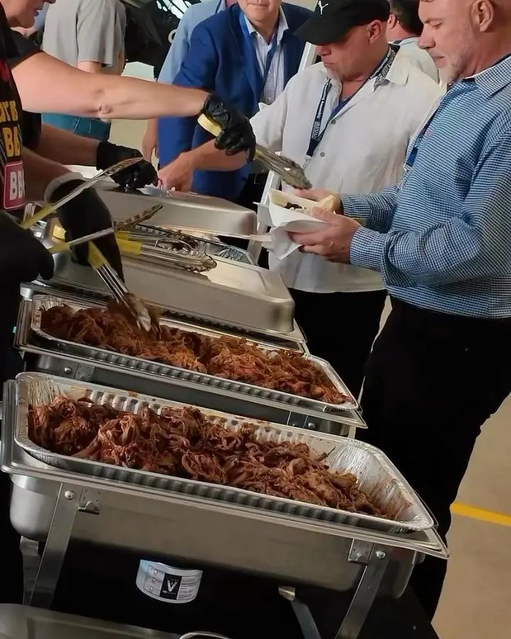 A company barbecue serve line: guests with plates moving along a row of chafing pans of pulled pork and brisket