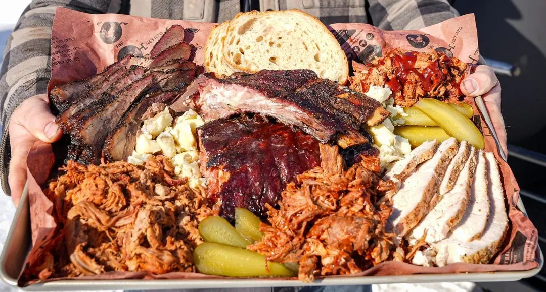 Close up of the tray: barked brisket, sauced ribs, piles of pulled pork, sliced chicken, macaroni salad, pickles and a thick slice of bread on printed butcher paper